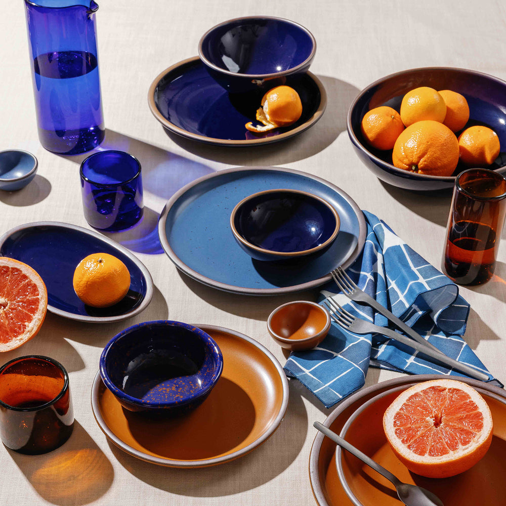 A styled tabletop dining scene with multiple ceramic plates and bowls in matte earthy orange and dark glossy blue, arranged with steel cutlery on a beige tablecloth. A blue-and-white check napkin, amber glass tumbler, oranges, and a halved grapefruit are also visible.