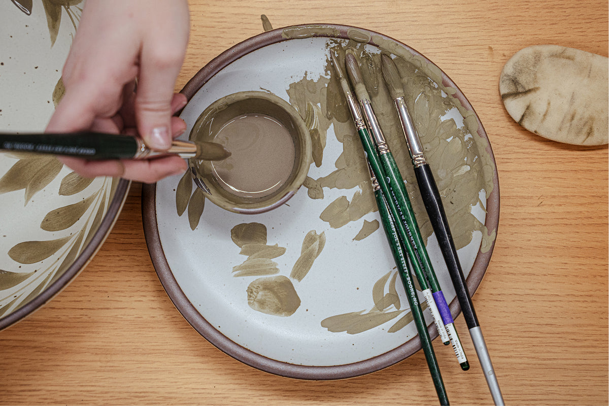 A hand holds a paintbrush over a ceramic plate being used as a palette on a wooden table, along with other paintbrushes.