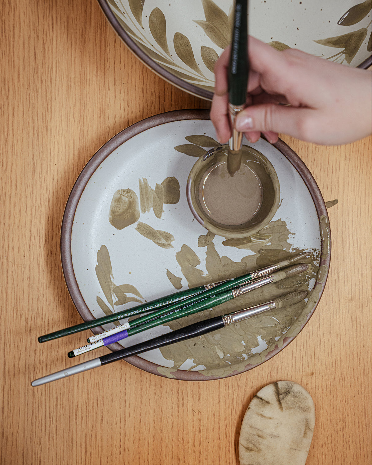 A hand holds a paintbrush over a ceramic plate being used as a palette on a wooden table, along with other paintbrushes.