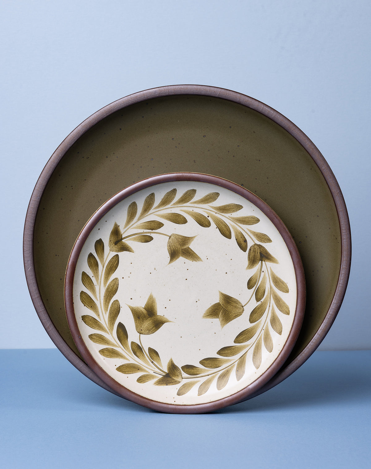 Two ceramic plates nested together, with a larger speckled olive green plate inside a smaller cream-colored plate decorated with a leaf wreath pattern, set against a light blue background.