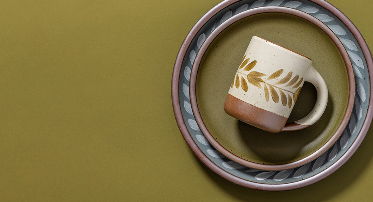 A ceramic mug with a leaf pattern on a cream and brown body, styled on a matching patterned plate against a solid olive green background.