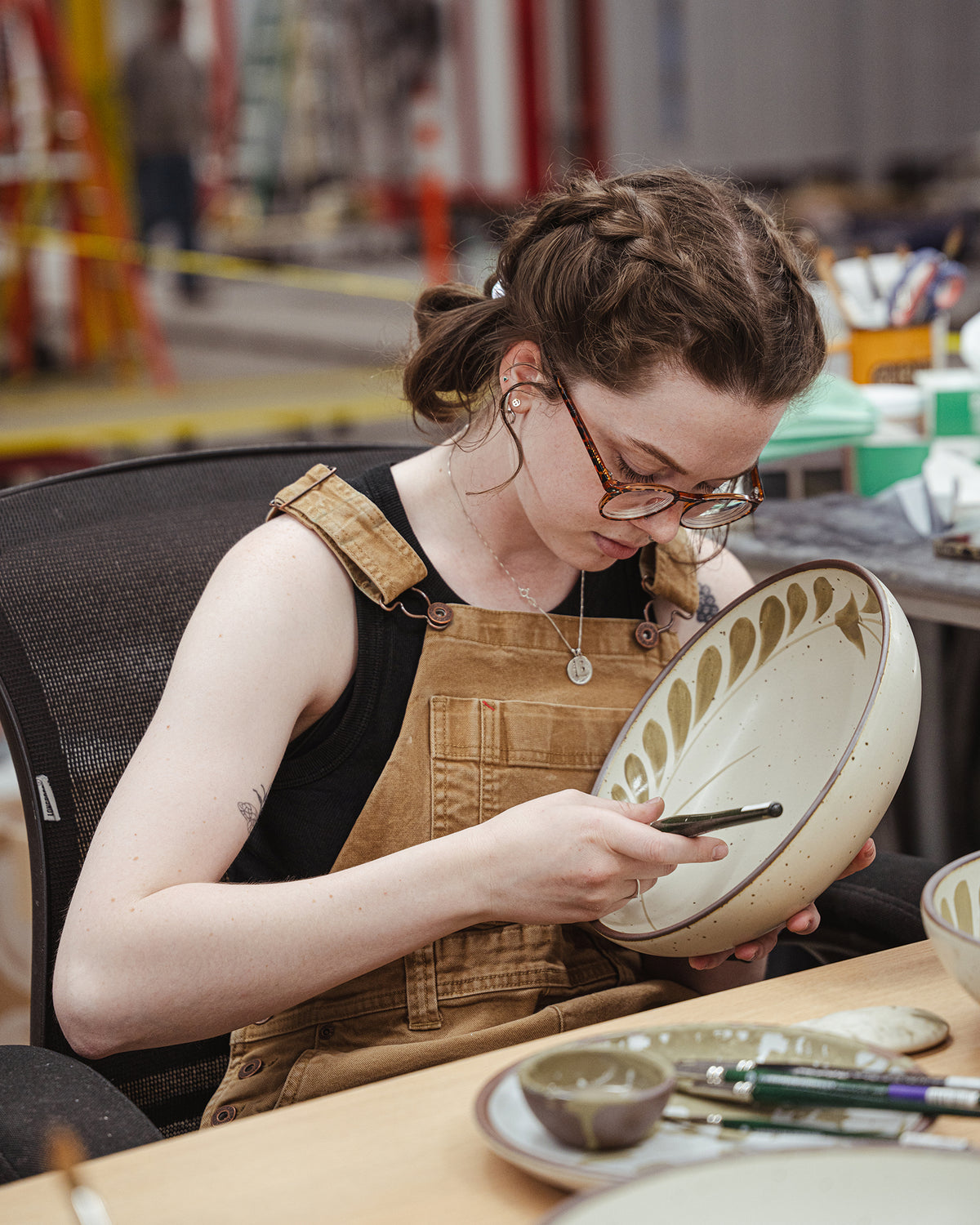 A person with glasses and braided hair sits at a table in a pottery studio, painting a hand-painted ceramic bowl with a paintbrush in hand.