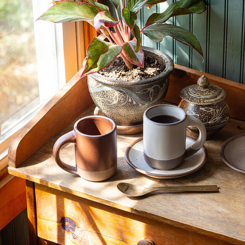 Two big mugs sitting on a side table with a spoon and potted plant.