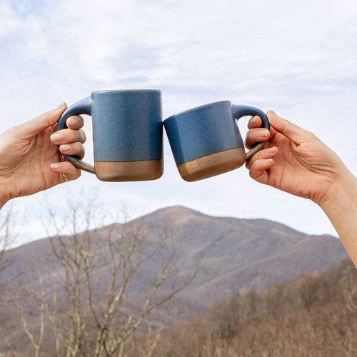 Outside, hands hold out The Mug and Small Mug in a grounded blue color with the mountains in the background.
