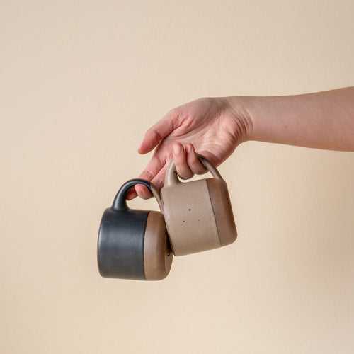A hand holds out a short ceramic mugs in a graphite black and pale warm brown color.