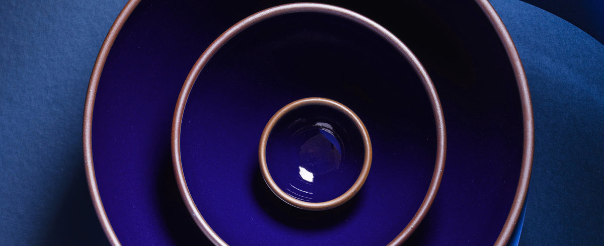 Three nested ceramic bowls in a glossy dark blue glaze with a contrasting brown rim, arranged concentrically on a dark blue background.