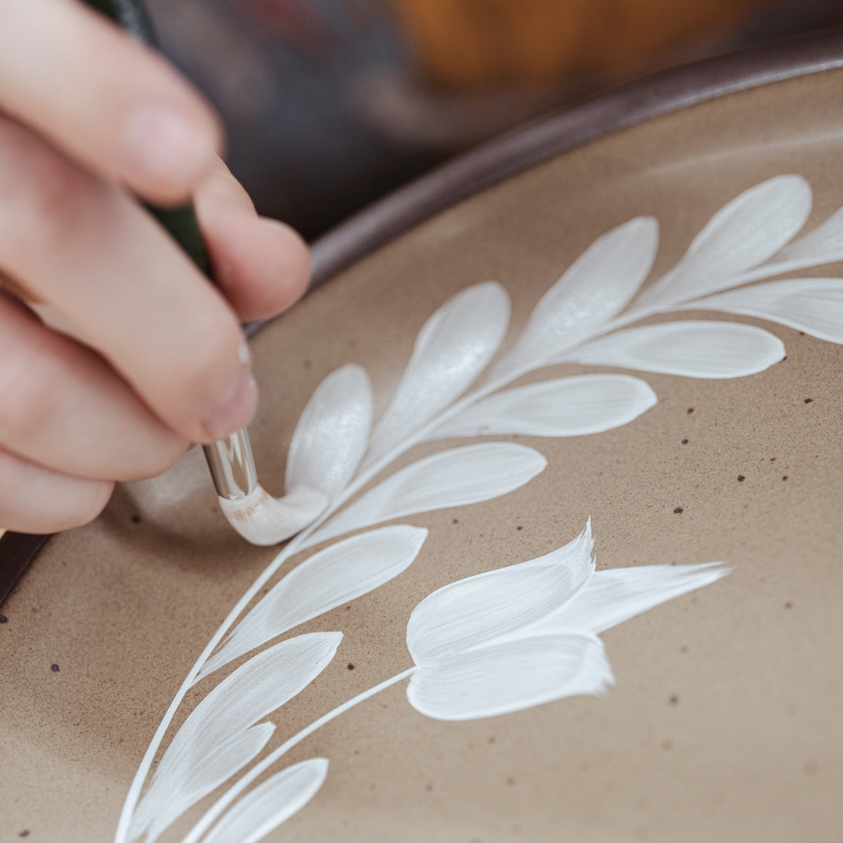 Hand painting a white greenery and tulip motif on a pale warm brown ceramic plate.