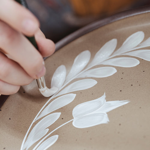 Hand painting a white greenery and tulip motif on a pale warm brown ceramic plate.