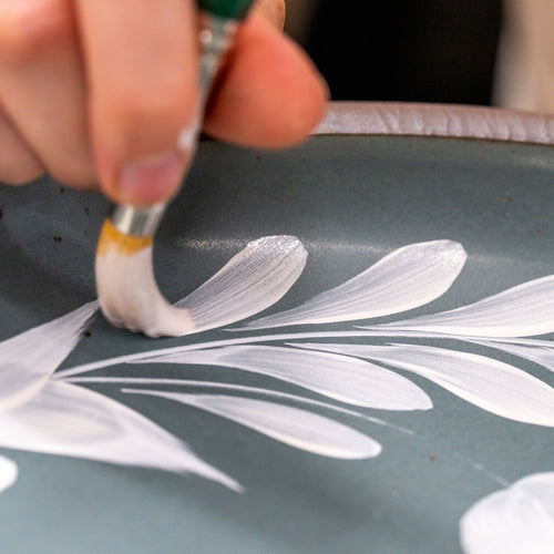 Hand painting a white greenery and tulip motif on a blue-grey ceramic plate.