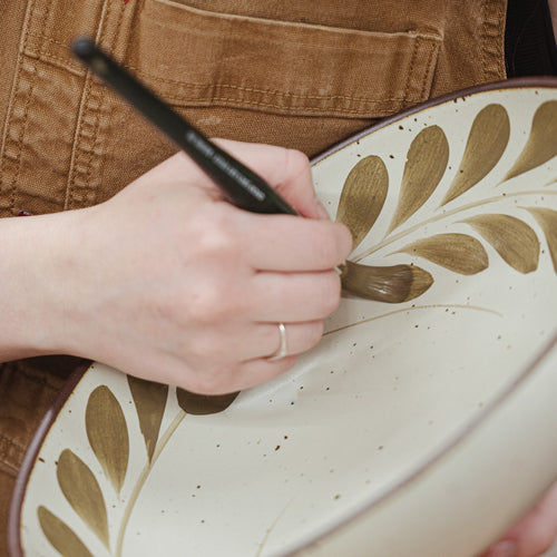 Hand painting an olive greenery and tulip motif on a cream ceramic plate.