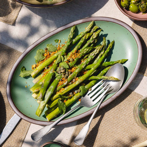 Green asparagus on a green medium oval plate with silverware on a textured surface