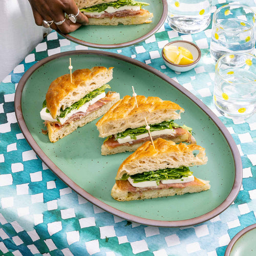 Plated sandwiches with toothpicks on a large green oval platter on a checkered tablecloth with drinks.