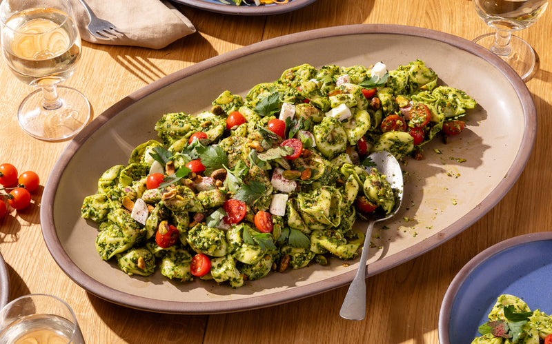 Family-style table setting with pesto pasta, colorful salad, and drinks served on oval ceramic platters and plates.