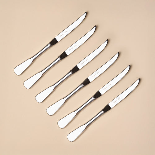 Six polished stainless steel dinner steak knives with rounded handles arranged diagonally on a neutral beige background.