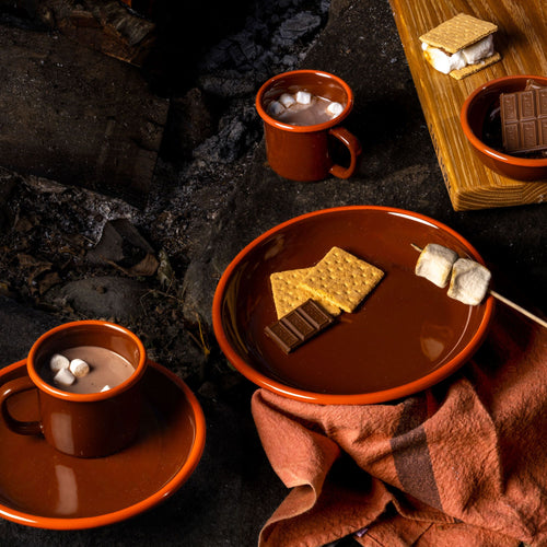 Enamel dinner plate in a red-brown color with smores along with enamel mugs filled with hot chocolate and marshmallows.