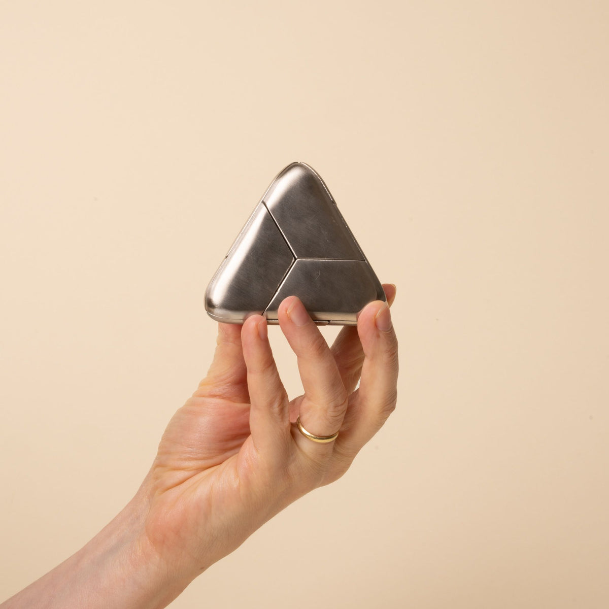 A hand holds a triangular silver metal puzzle with smooth rounded edges and subtle seam lines, resting on a beige background.