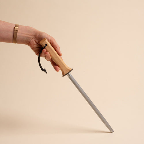 Hand holding a honing steel with a wooden handle and black loop against a neutral beige background.