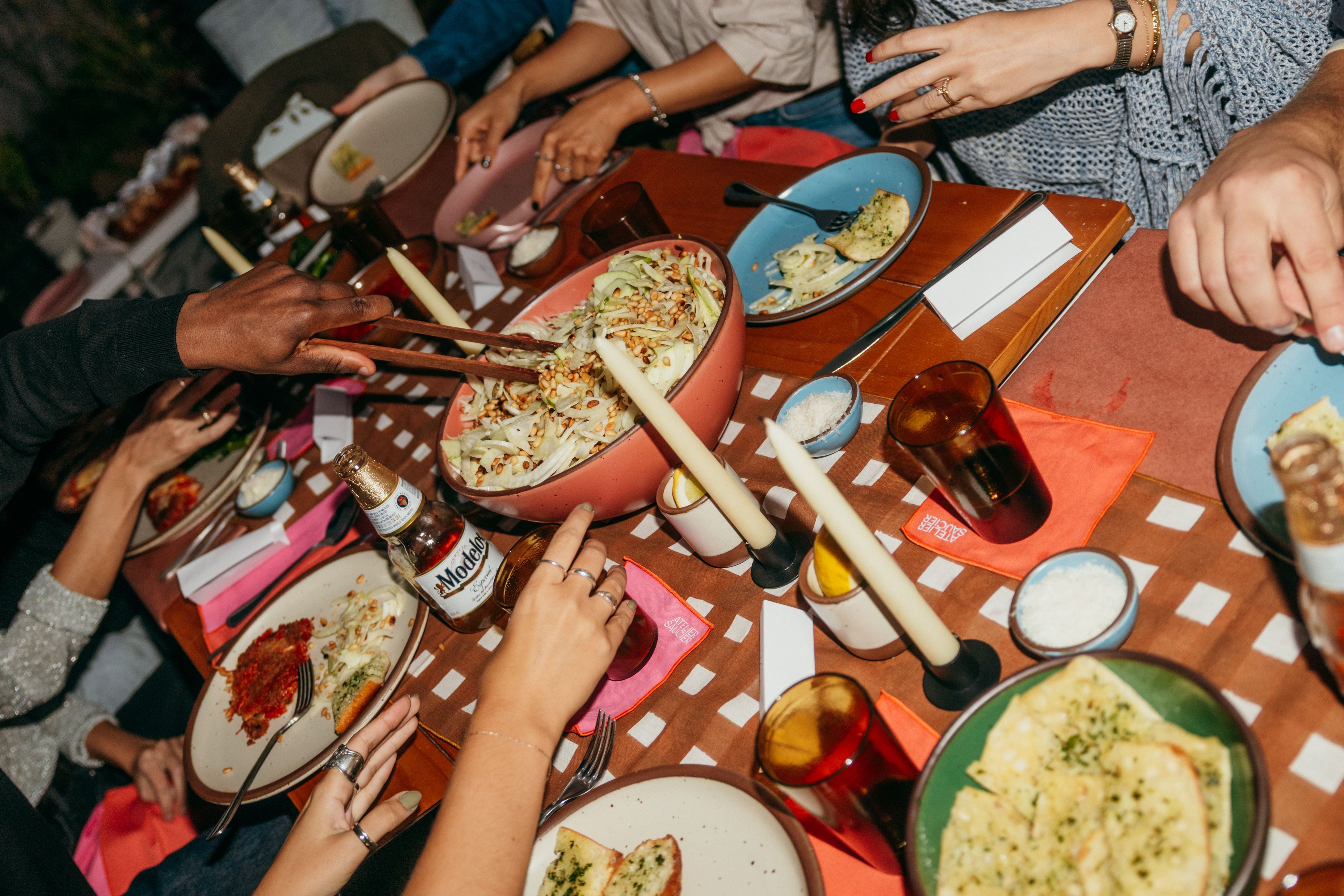 A table set with plates and bowls of food, glassware and taper candles with hands reaching for different items.