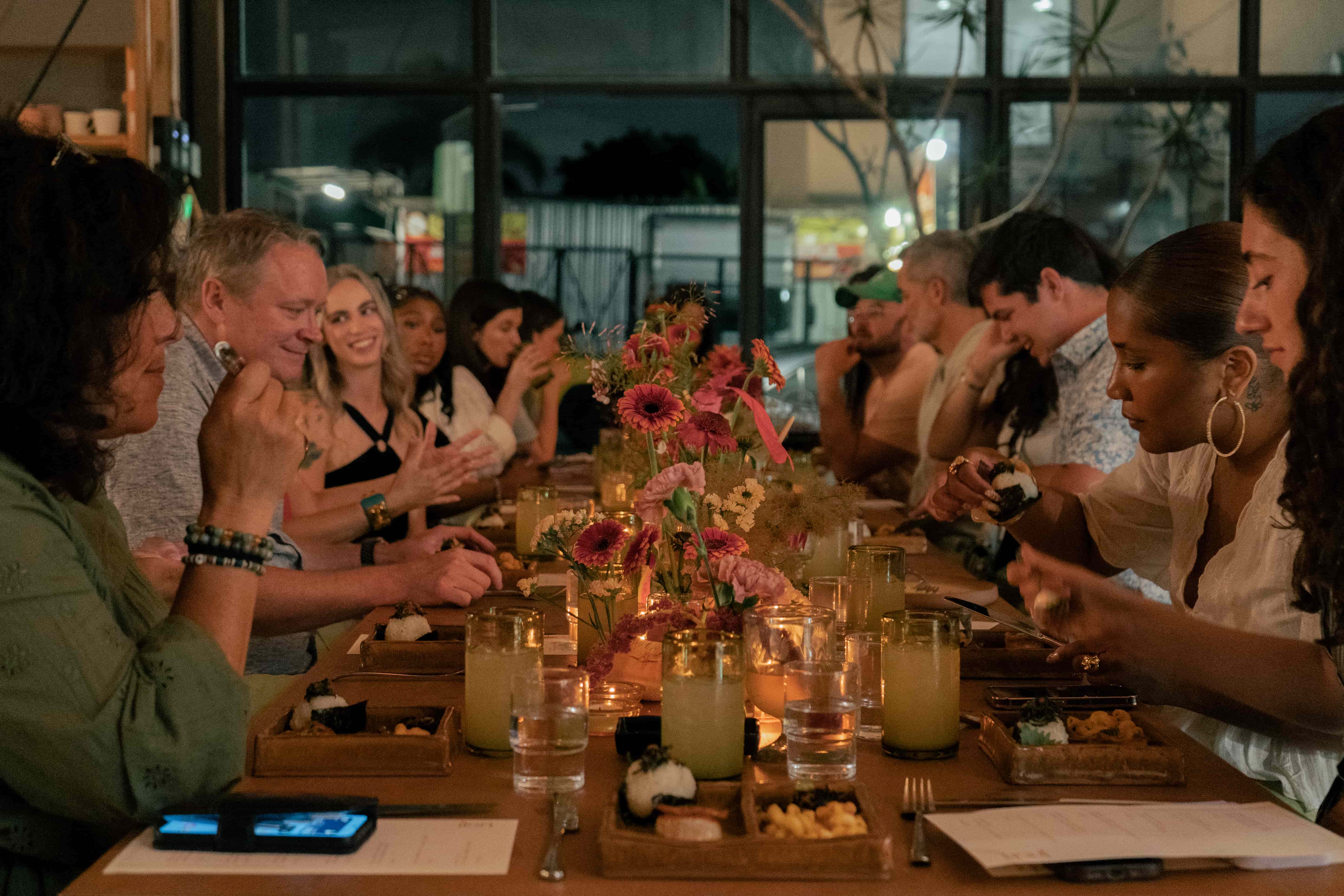 A large table filled with people sitting down eating and talking, the table is filled with menus, flowers, glasses, and moody candlelight.