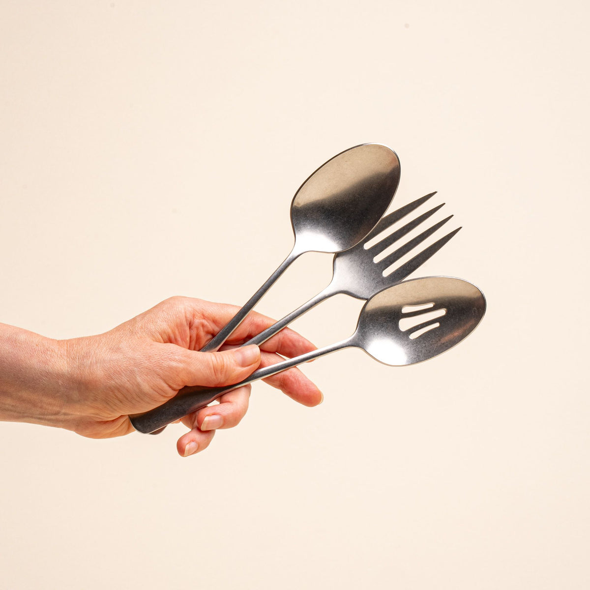 A hand holds three stainless steel serving utensils including a serving fork, serving spoon, and pierced spoon in a tumbled matte finish with a classic design.