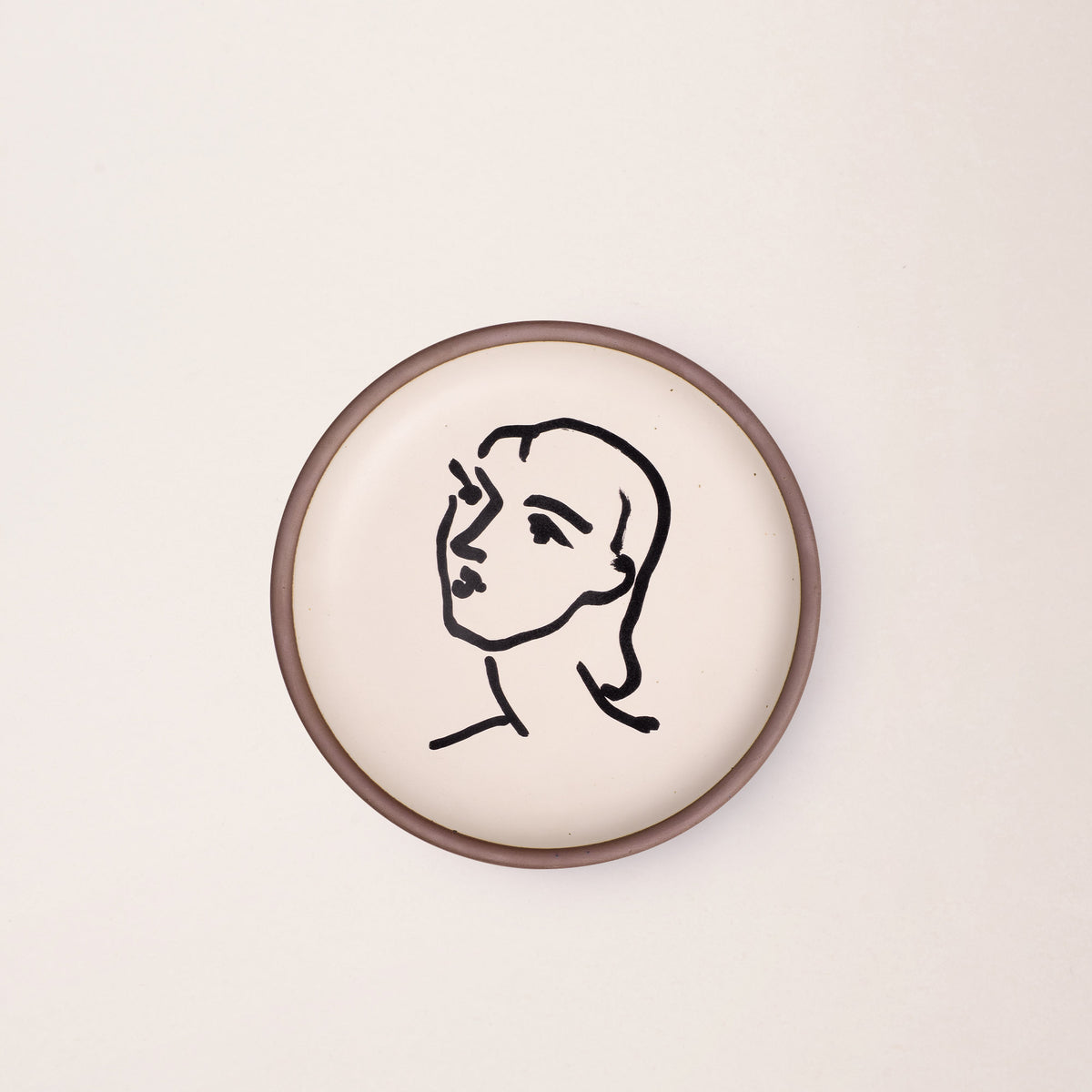 A ceramic plate featuring a decal of the artwork, Nadia aux cheveux lisses, by Henri Matisse. A minimalist black ink decal of a woman's face and neck in profile, facing left, in an abstract, expressive style centered on an off-white ceramic plate with an unglazed rim.