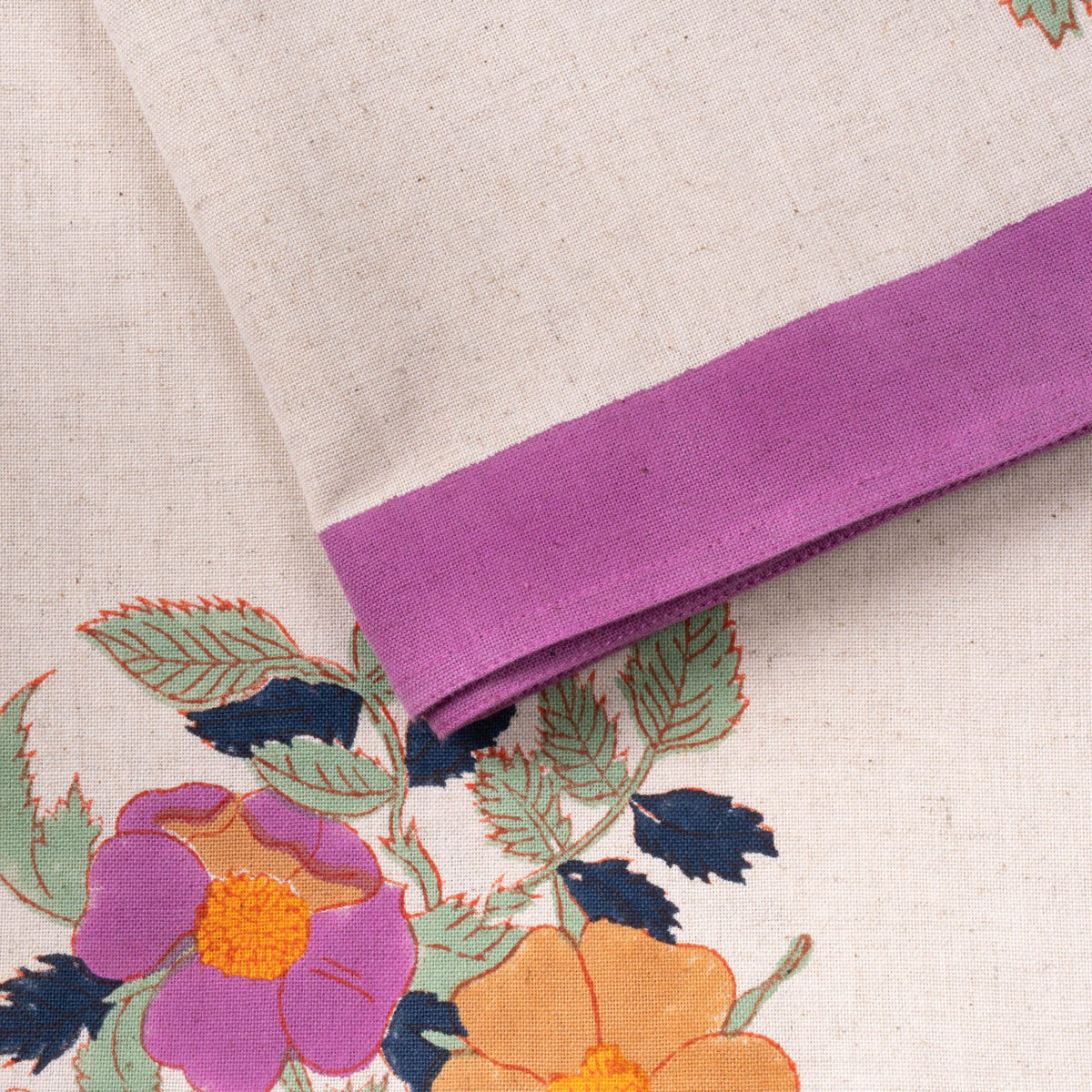 Close-up of a cream linen tablecloth with a purple border and a floral print in purple and orange with green leaves.
