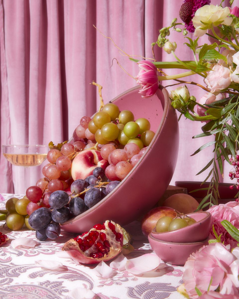 A large pink bowl is tipped on its side and filled with varying colors of grapes and other fruits. A flower arrangement, pink fabric background, and champagne glass is surrounding the bowl.