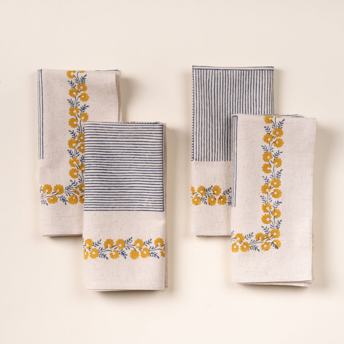A set of four folded cream-colored napkins with blue striped accents and yellow floral border detailing, arranged neatly on a light background.