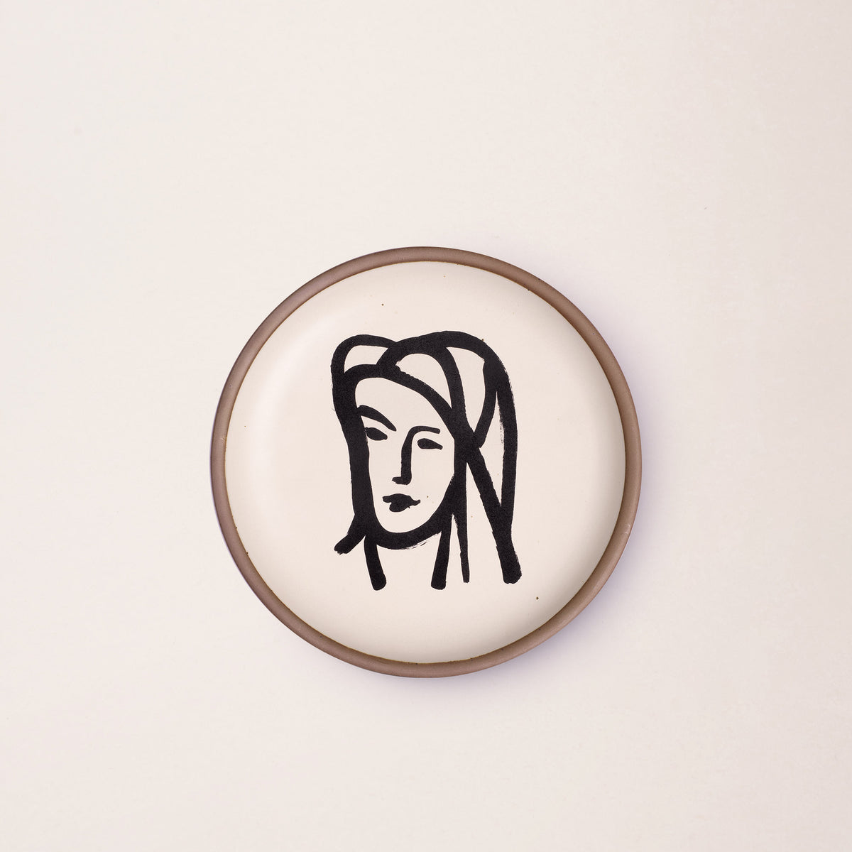 A ceramic plate featuring a decal of the artwork, Bédouine au grand voile, by Henri Matisse. A minimalist black ink decal of a woman's face wearing a large veil depicted by bold brushstrokes in an abstract, expressive style centered on an off-white ceramic plate with an unglazed rim.