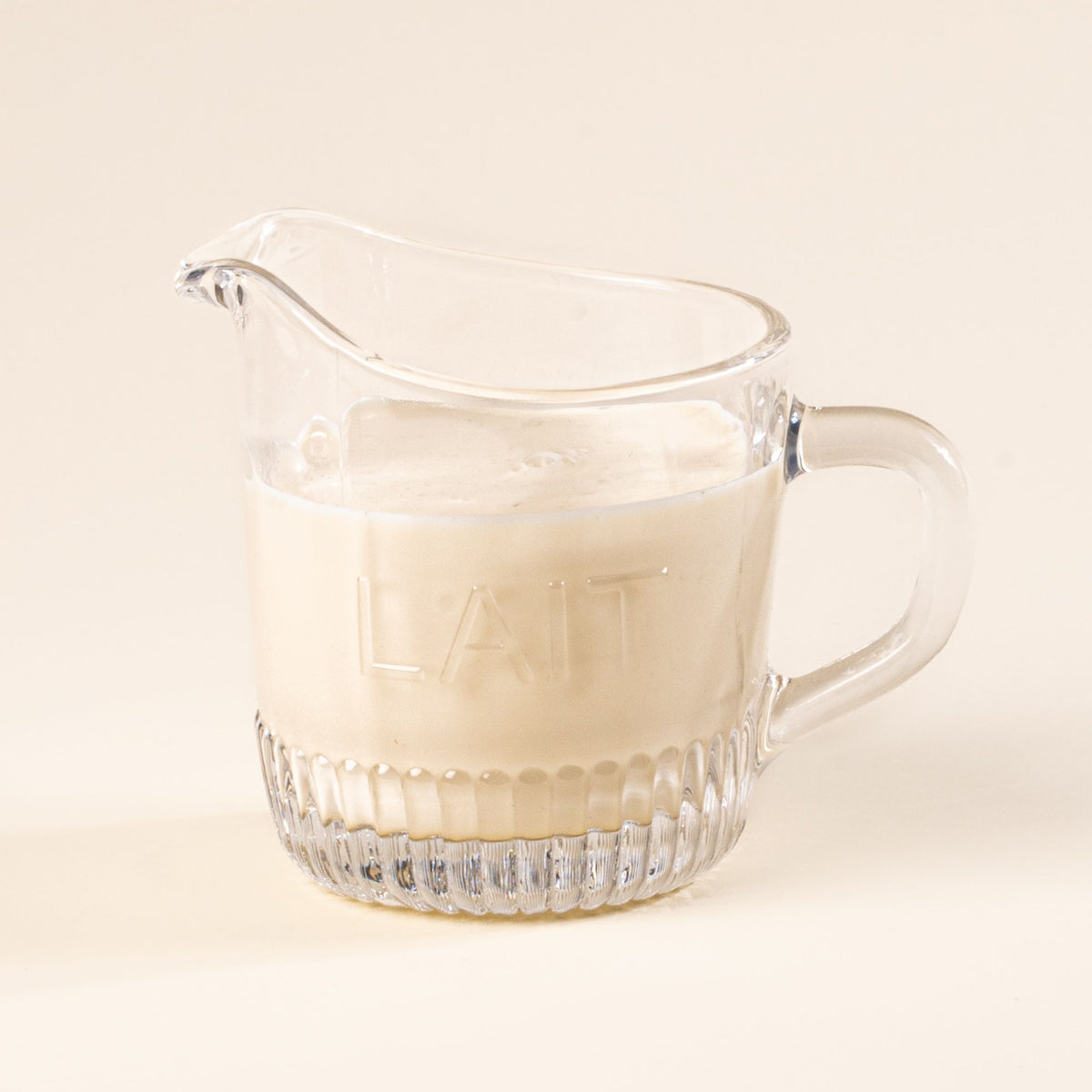 Clear glass pitcher with handle and spout on a light background, filled with milk