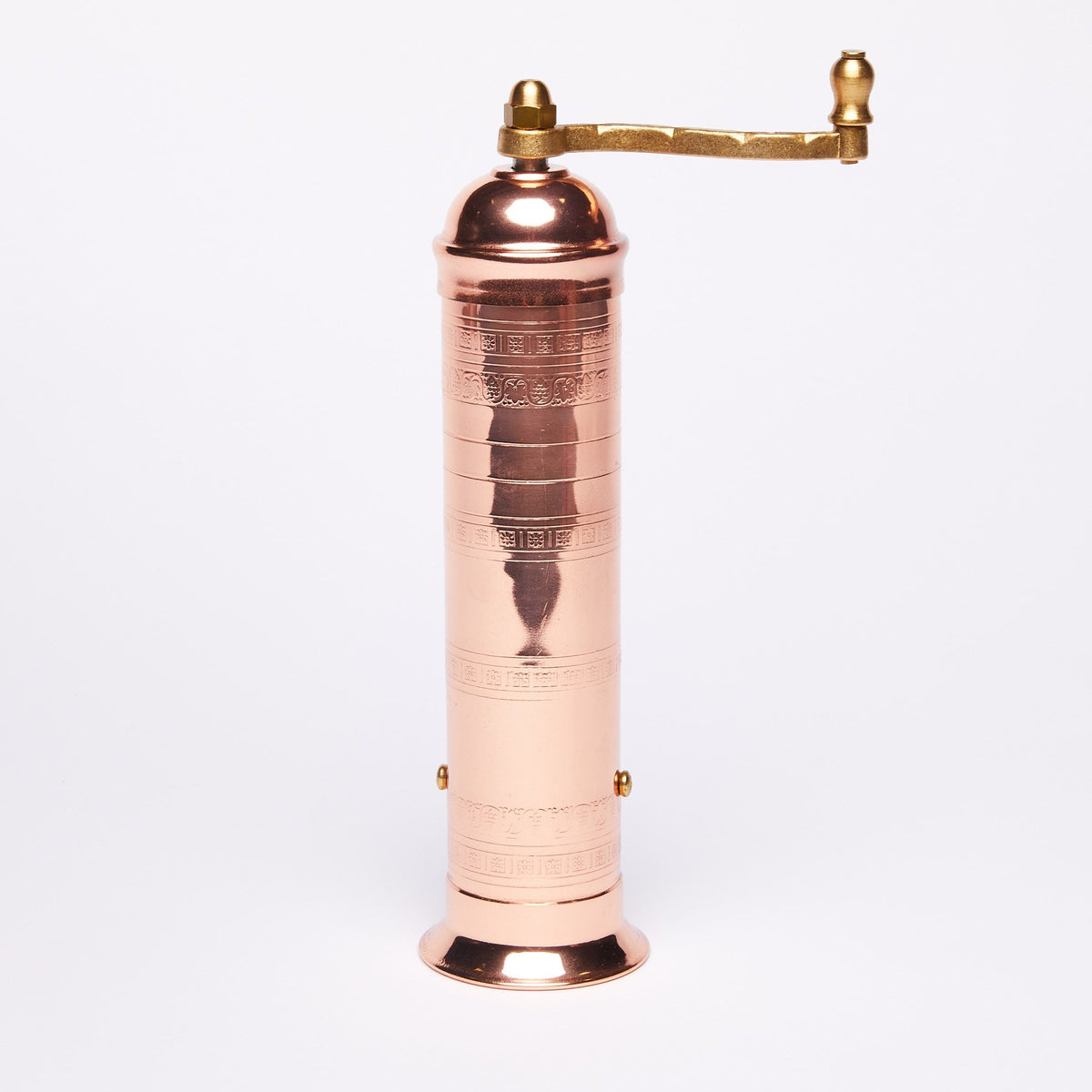 Copper manual pepper mill with a tall cylindrical body and intricate engraved patterns, featuring a brass handle on top, set against a plain white background.