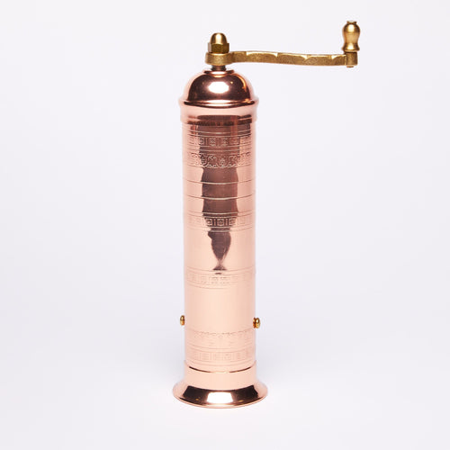 Copper manual pepper mill with a tall cylindrical body and intricate engraved patterns, featuring a brass handle on top, set against a plain white background.