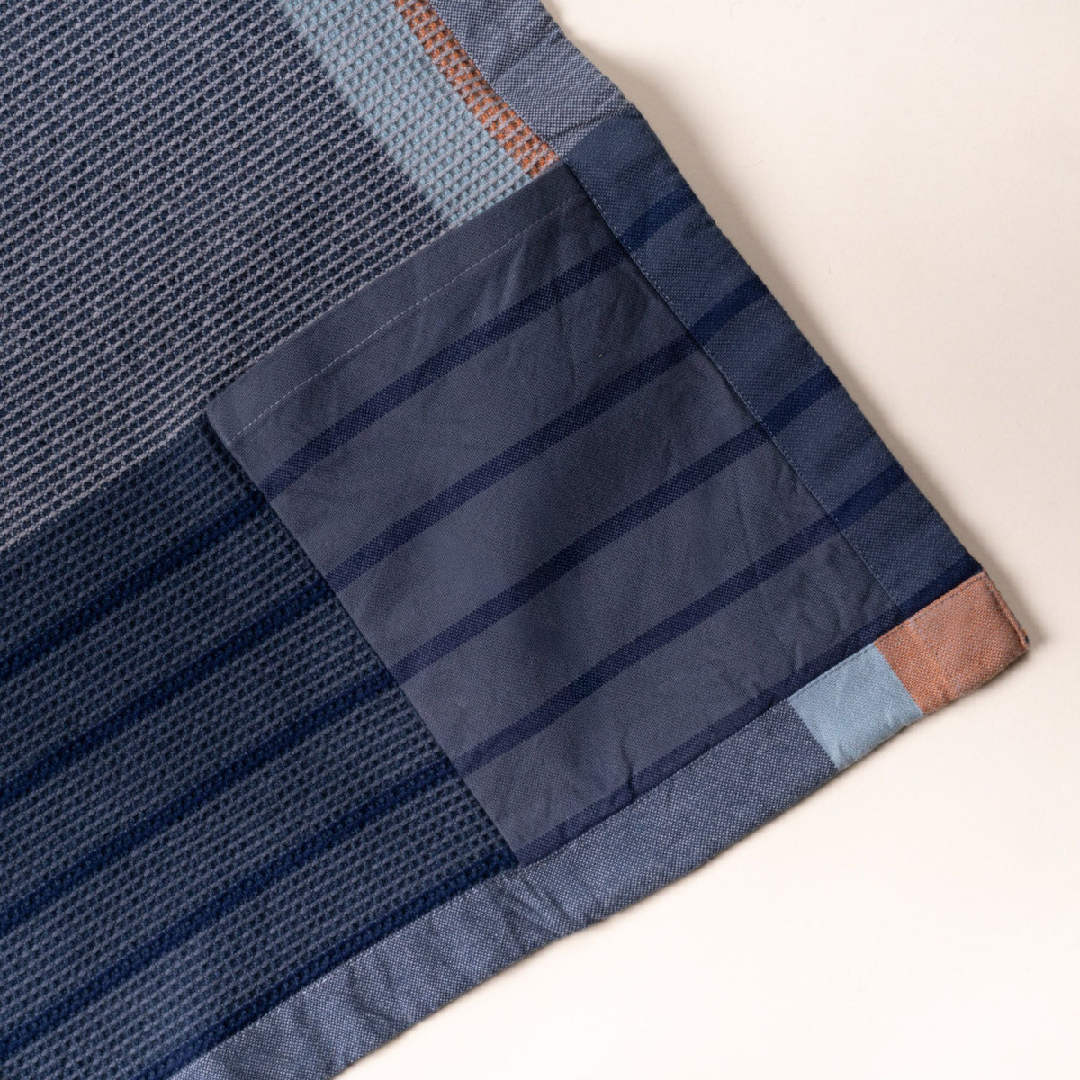 Close-up of a navy and slate-blue woven apron corner, showing a striped front pocket and trim with small light blue and rust accent blocks.