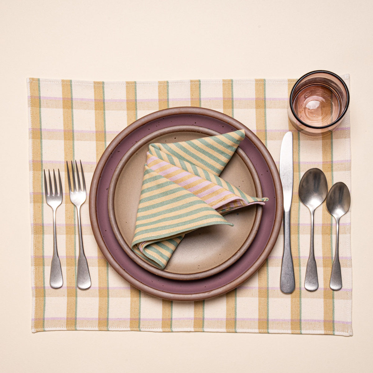 A place setting featuring ceramic plates in warm pale brown and soft mauve purple colors, a folded striped napkin, silverware, and a glass, on a pastel plaid placemat.