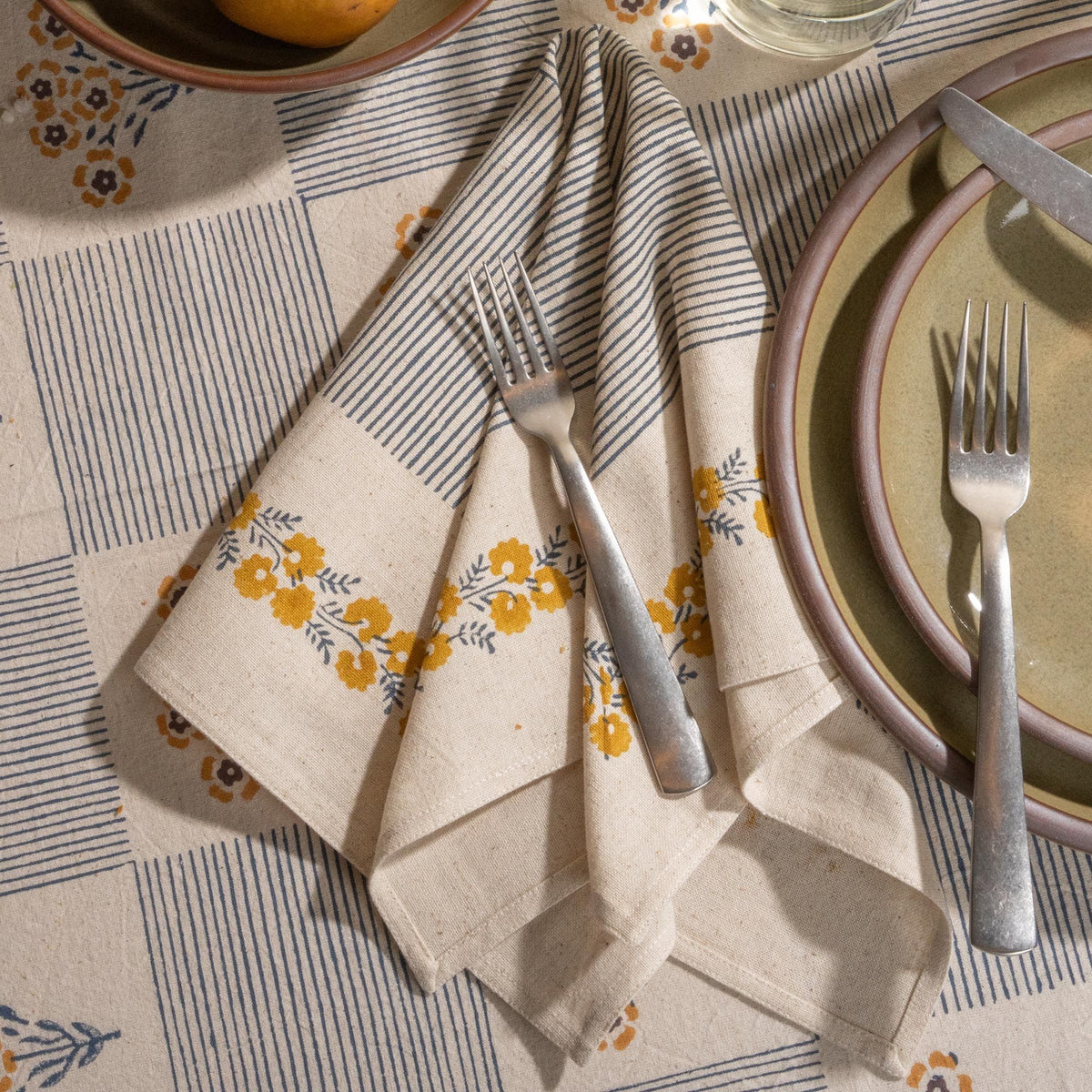A cream-colored napkins with blue striped accents and yellow floral border detailing, arranged with silverware next to grey-green ceramic plates.