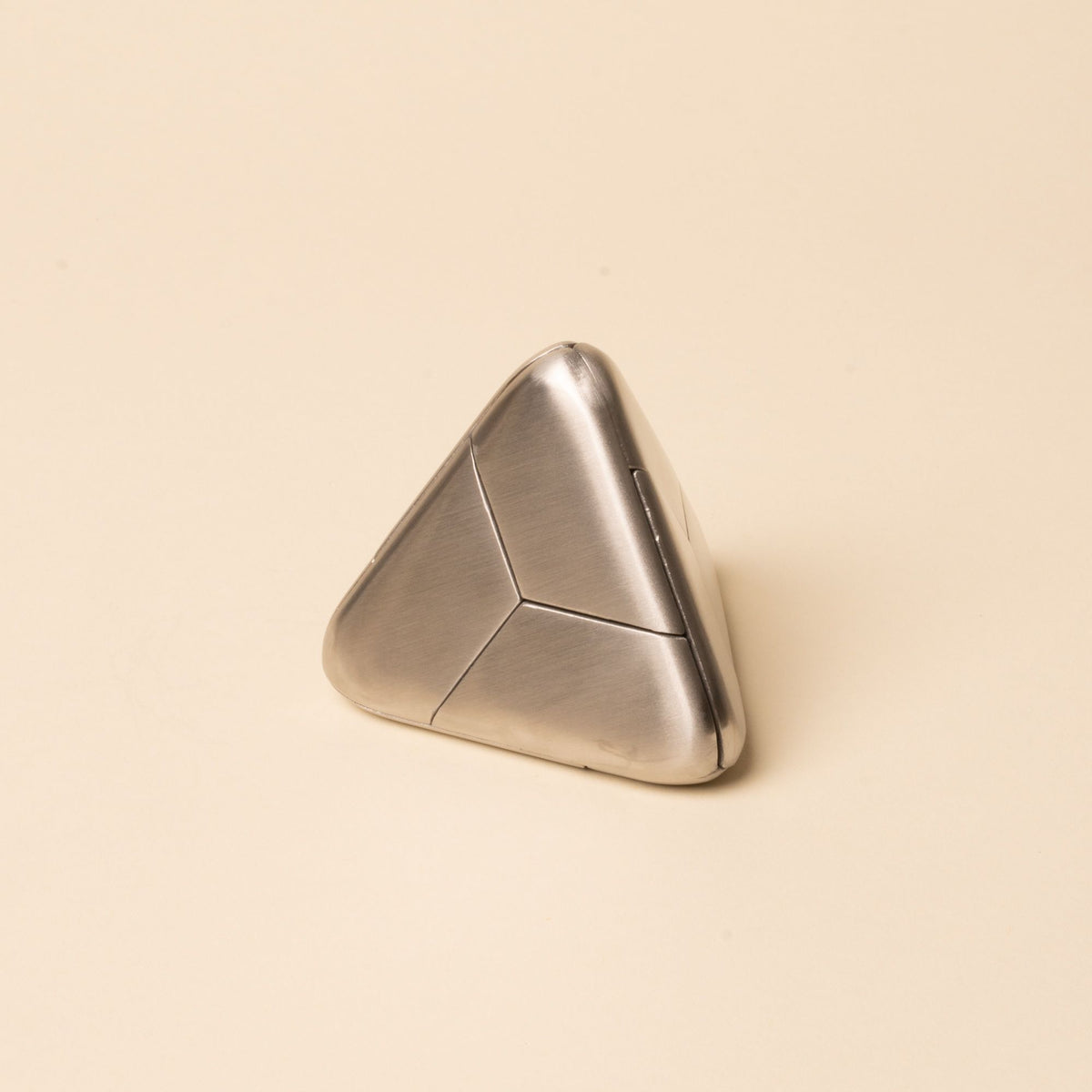 Triangular silver metal puzzle with smooth rounded edges and subtle seam lines, resting on a beige background.