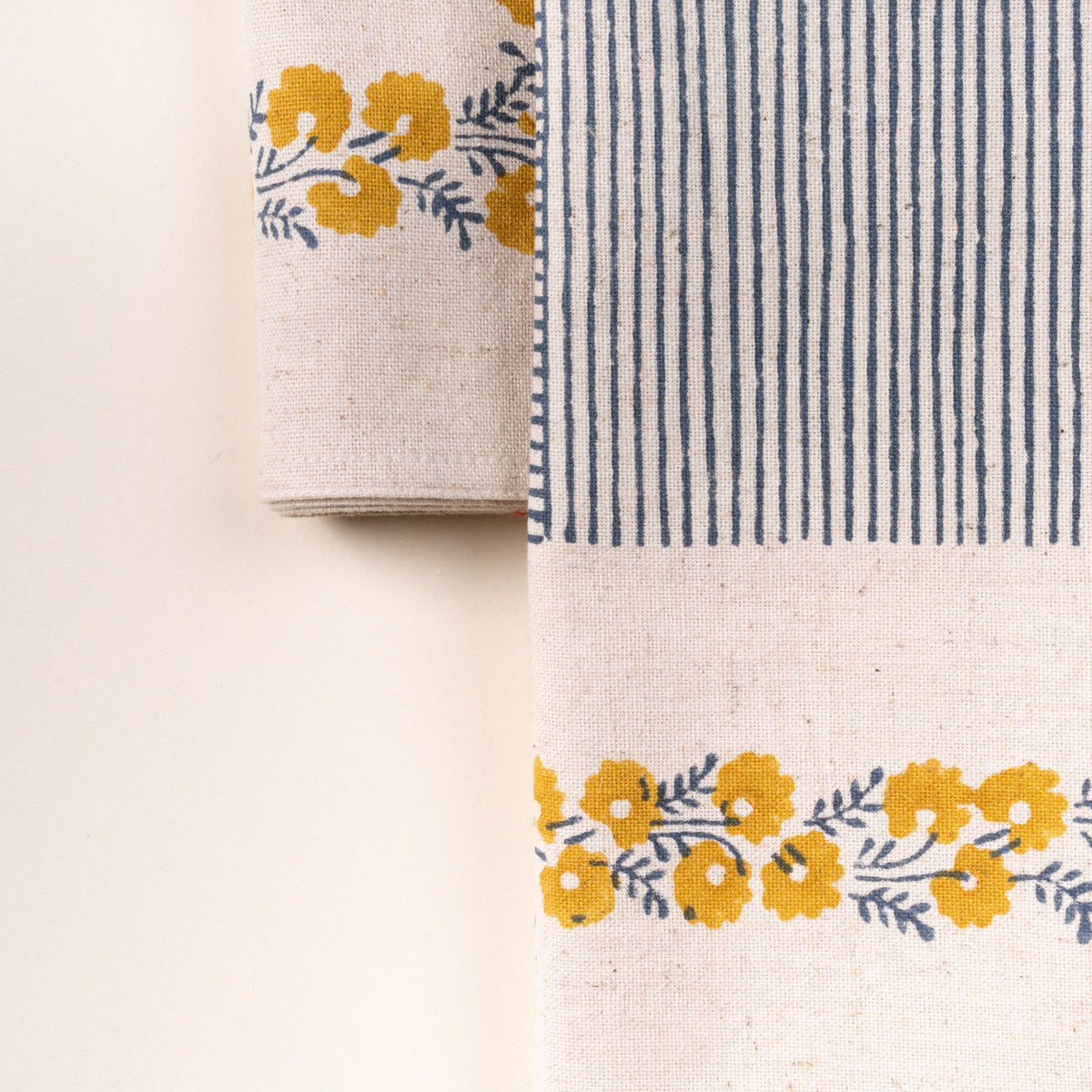 A closeup of a cream-colored napkin with a delicate yellow floral border framing a central blue striped patchwork design, laid flat against a neutral background.
