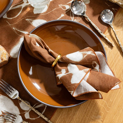 Glossy caramel ceramic plate with patterned napkin in a gold ring, set on a table with matching linens and sand drip flatware.