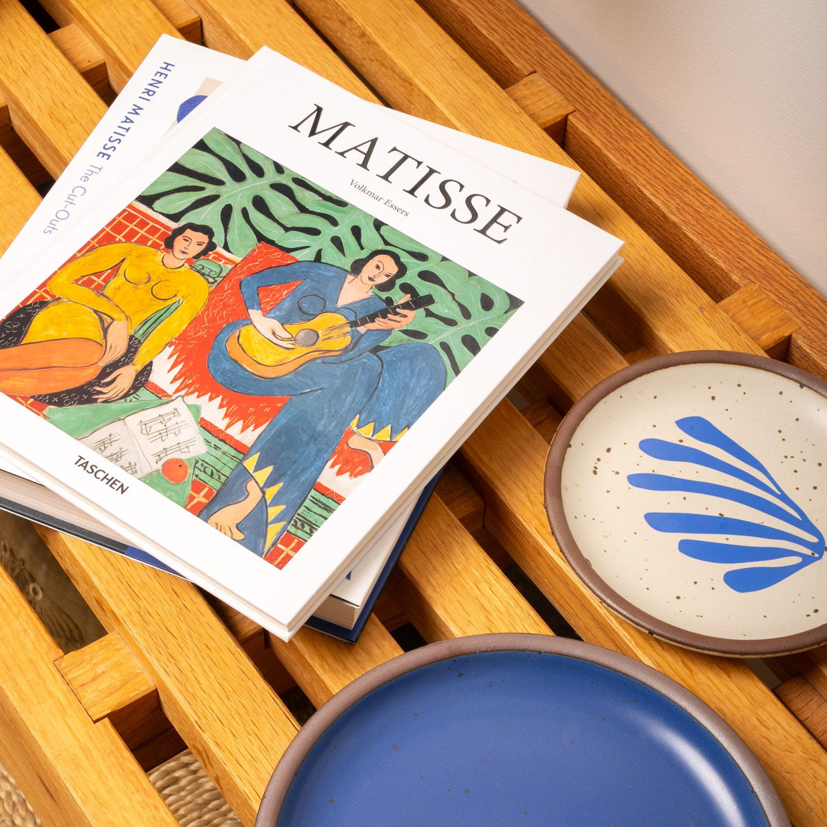 Two art books on Matisse rest on a wooden slatted bench beside ceramic plates in blue and cream with bold blue brushstroke designs.
