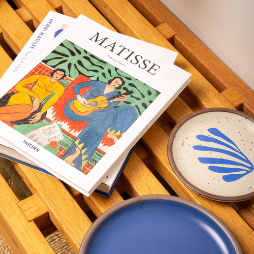 Two art books on Matisse rest on a wooden slatted bench beside ceramic plates in blue and cream with bold blue brushstroke designs.