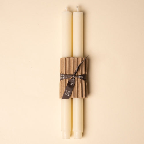 Two ivory taper candles bundled with corrugated cardboard and tied with a black ribbon on a neutral background.