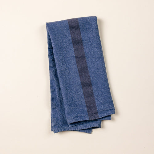 A folded linen towel in an indigo color with a plum stripe in the middle