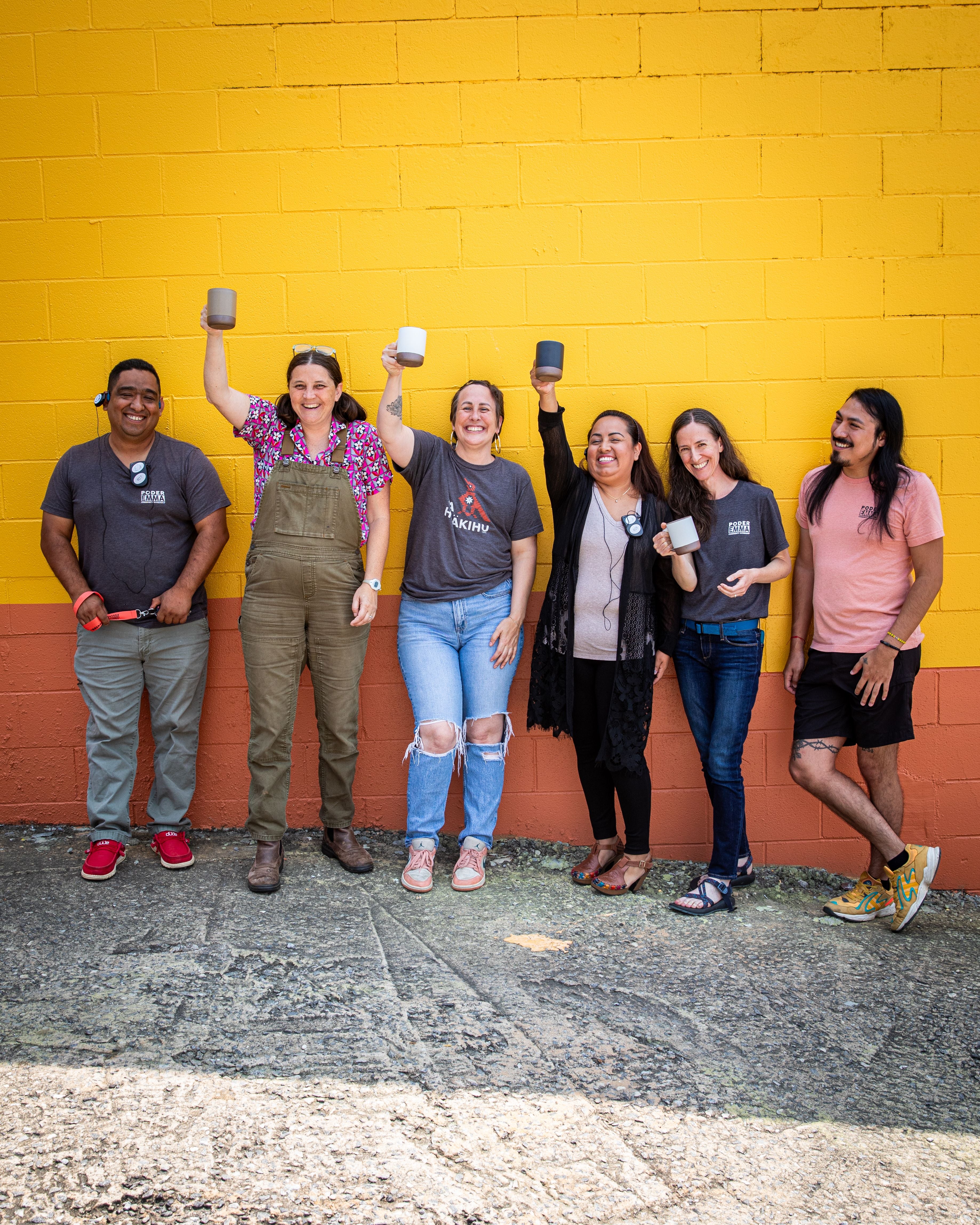 Six members of PODER Emma stading in front of a yellow and orange wall holding mugs and smiling