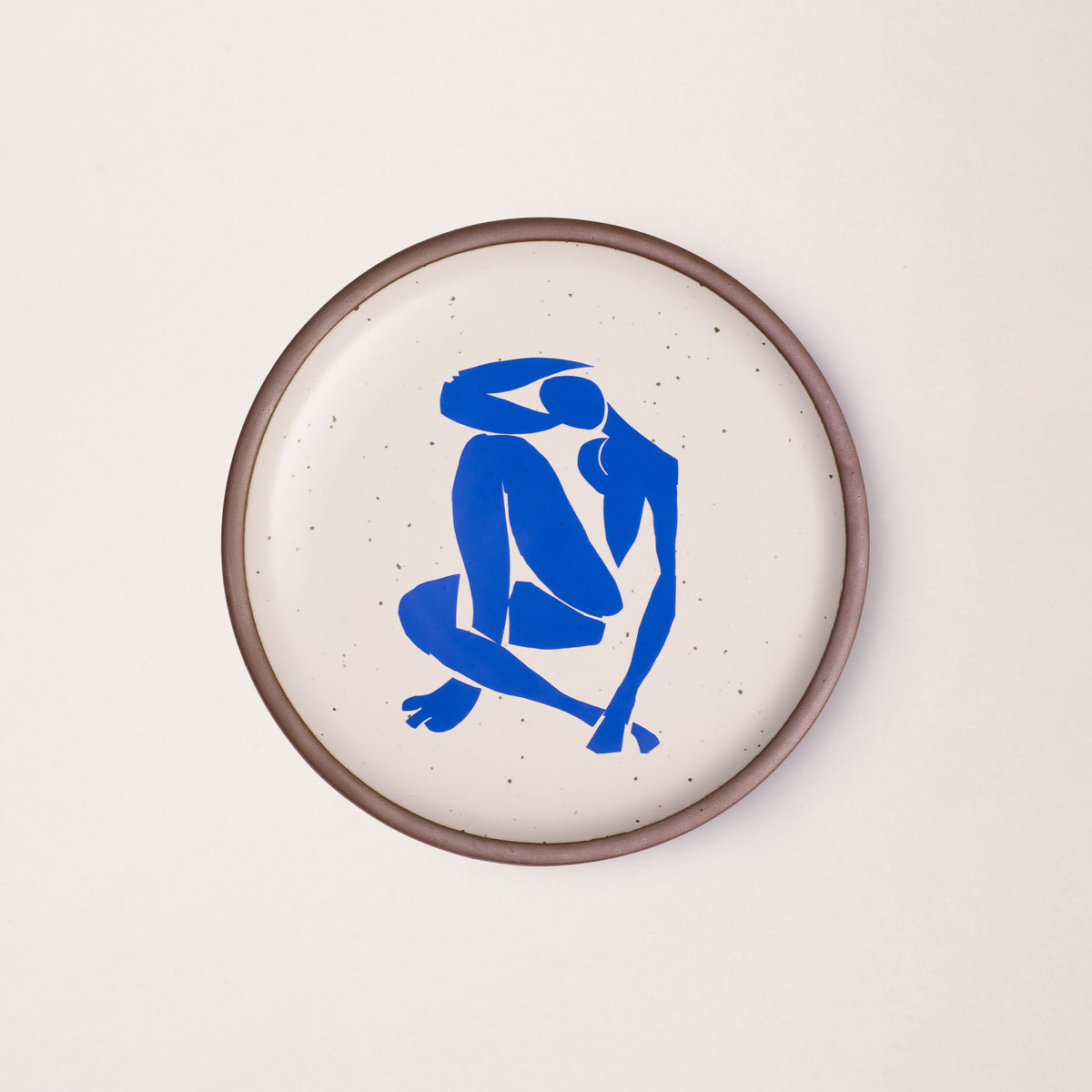 A plate featuring Nu Bleu IV, by Henri Matisse, a bright blue, abstract, silhouetted female figure in a curled, seated position, with sharp cut-out edges, centered on an off-white ceramic plate with an unglazed rim.