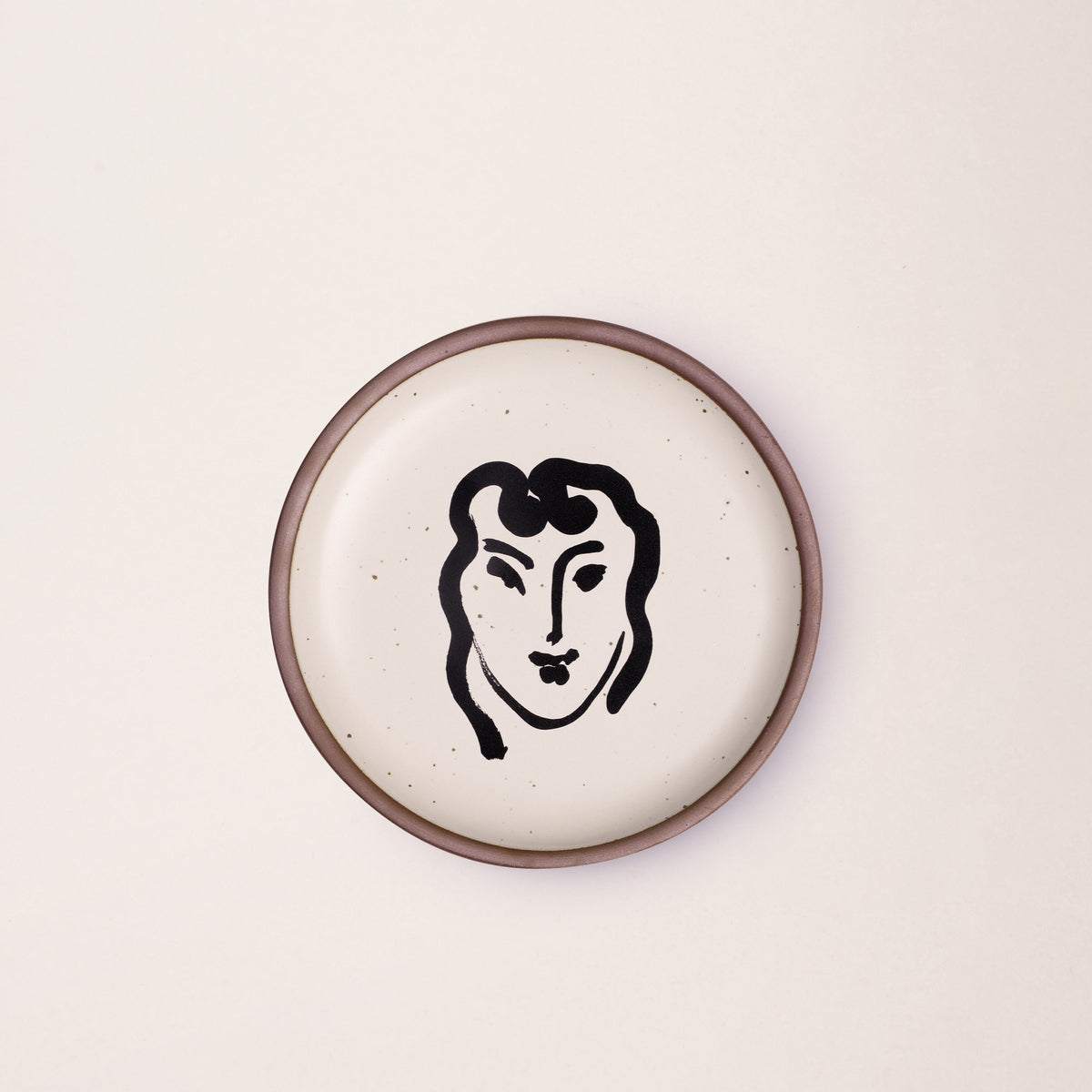 A ceramic plate featuring a decal of the artwork, Patitcha souriante, by Henri Matisse. A minimalist black ink decal of a woman's smiling face in an abstract, expressive style centered on an off-white ceramic plate with an unglazed rim.