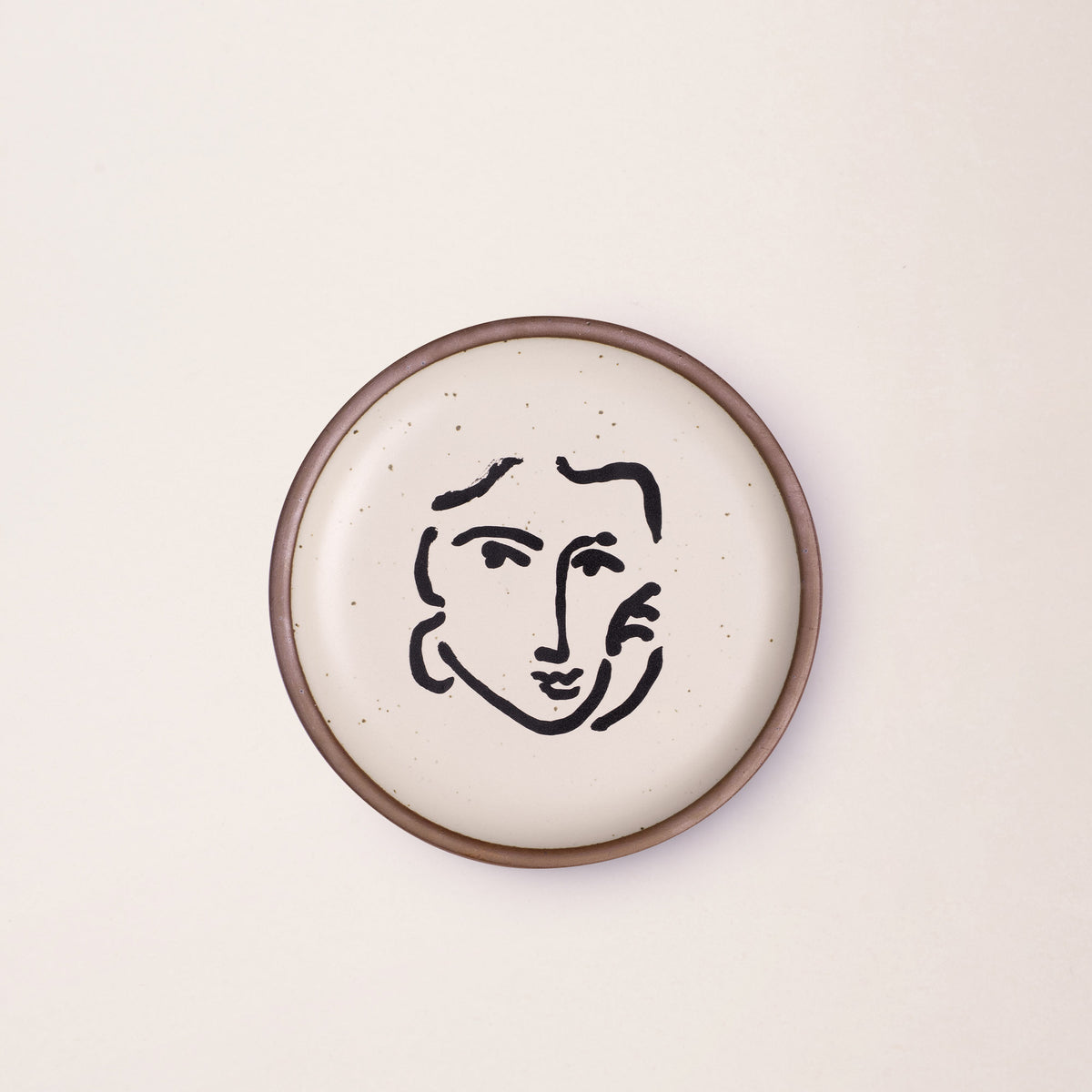 A ceramic plate featuring a decal of the artwork, Nadia au regard sérieux, by Henri Matisse. A minimalist black ink decal of a woman's face and hand resting on one cheek, in an abstract, expressive style centered on an off-white ceramic plate with an unglazed rim.