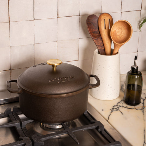Black cast iron Dutch oven with brass knob on stovetop, beside ceramic utensil holder with wooden spoons and olive oil bottle.