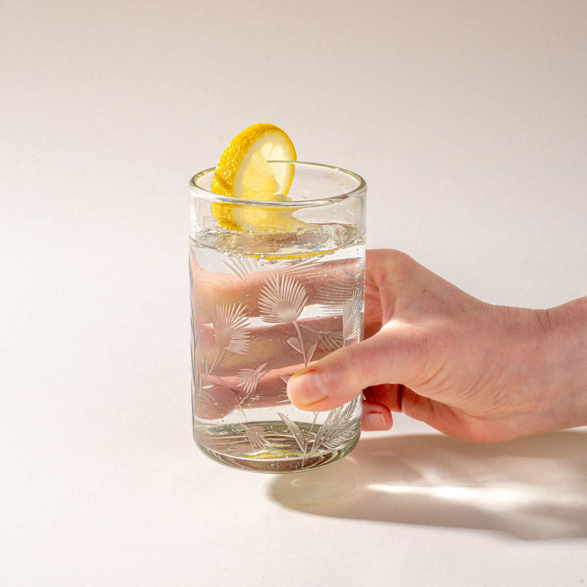 A hand holds a clear tumbler glass made of recycled glass with etched thistle designs on it filled with water and topped with a slice of lemon.