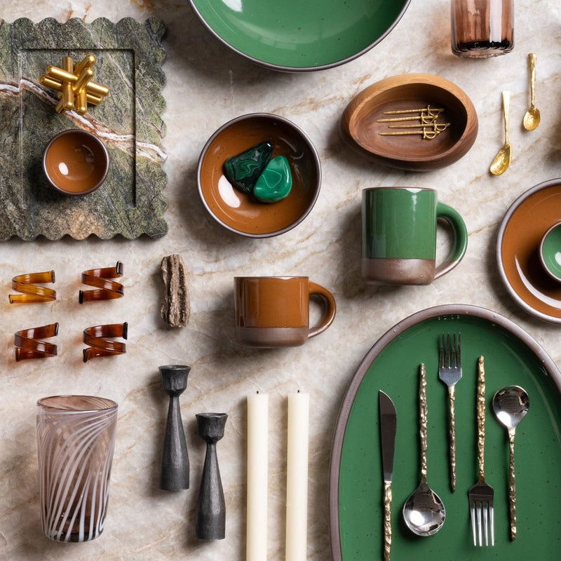 A collection of ceramic tableware in green and brown glazes, including mugs, bowls, and a large oval serving platter with utensils. The setting also features candle holders, candles, a tumbler, and decorative items on a marble surface.
