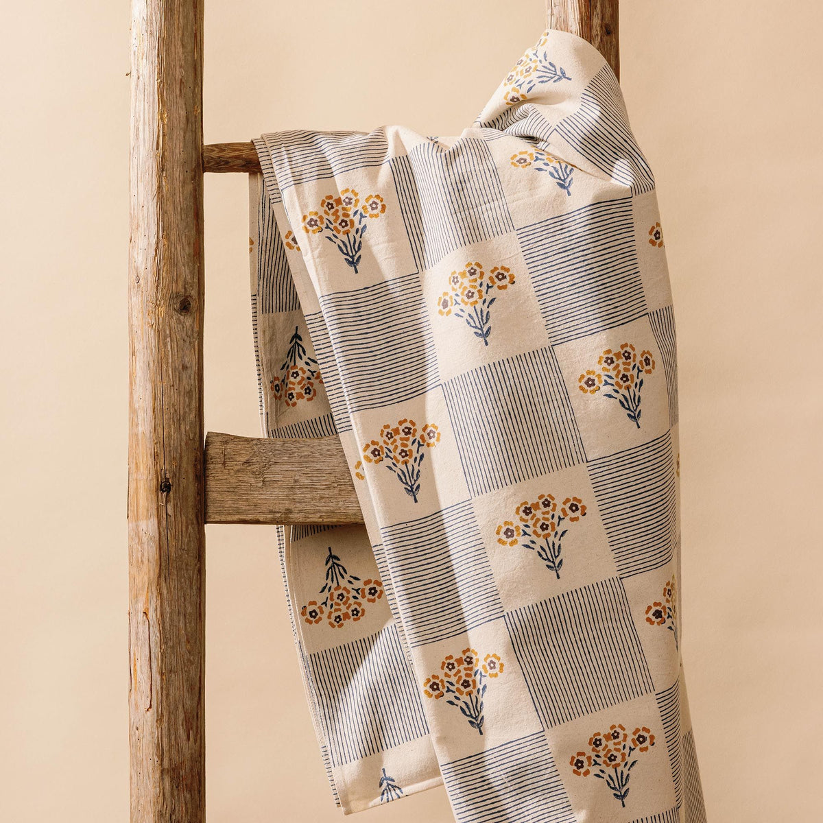Folded blue-and-white checkered cloth tablecloth printed with small yellow flower bouquets on a wooden ladder.